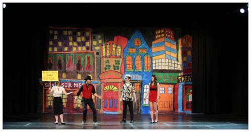 (Image: A Missionary Holds a Sign Advertising a
 Prayer Meeting while Other Cast Members Discuss the
 Situation)