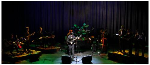 (Image: Orange and Green Bathe the Band while
 Wisps of Light Illuminate the Rear Curtain)