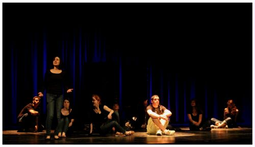 (Image: Actors sit in Various Positions
 around the Stage while one Singer Stands)
